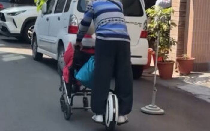 Người đàn ông lớn tuổi cưỡi xe điện một bánh, vừa chạy vừa đẩy xe lăn trên đường