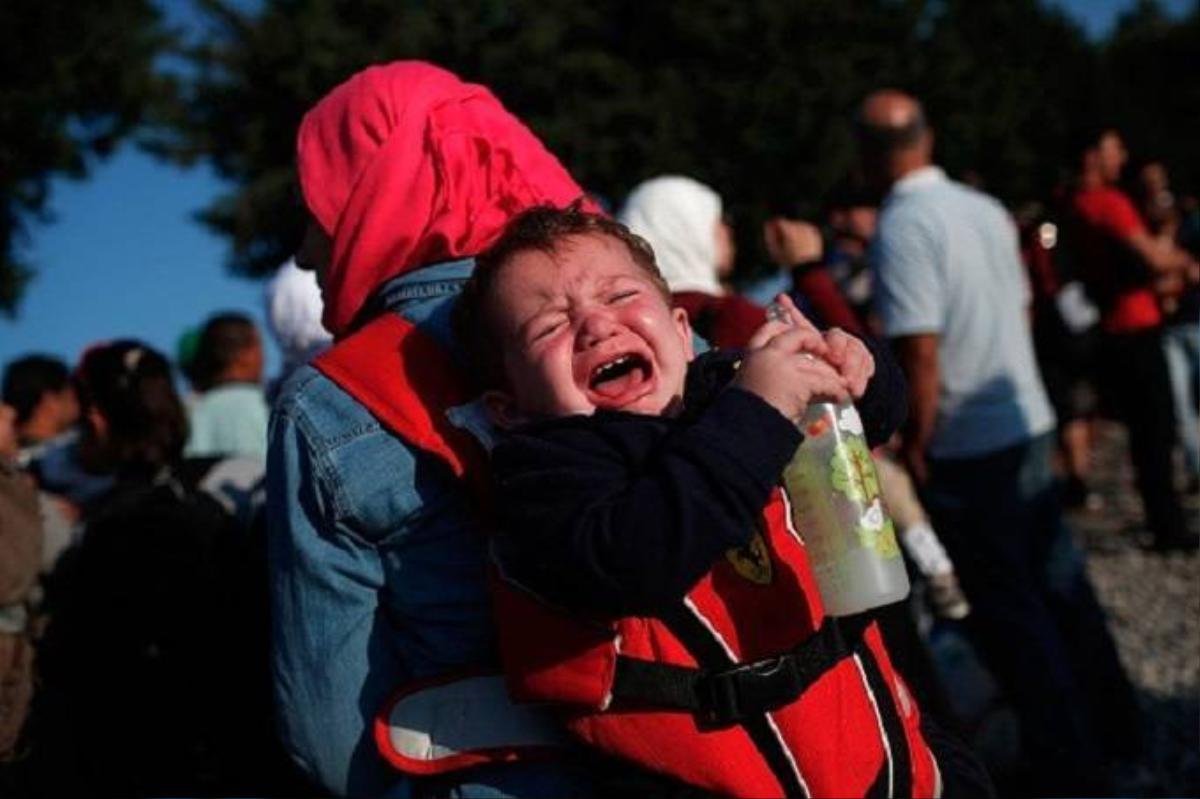 Em bé Syria khóc trên lưng mẹ khi hai mẹ con đang đợi tàu ở thành phố Idomeni, Hy Lạp. Từ đây, người di cư sẽ tiếp tục tiến về hướng bắc để tới Macedonia.