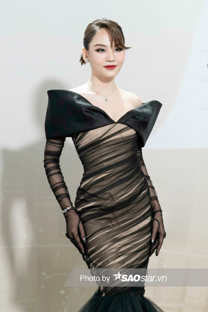 Hoa hậu Mai Phương tựa thiên nga đen với thiết kế trễ vai được làm từ chất liệu voan. Người đẹp sinh năm 1999 buộc gọn tóc để lộ phần mái hờ hững làm tăng thêm phần mới mẻ cho tổng thể. 