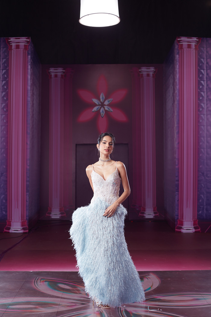 Nối gót Trương Quỳnh Anh, người mẫu Emma Lê sải bước tự tin trong thiết kế hai dây xẻ ngực chữ V có màu bạc kết hợp lông vũ xanh dương. Chiếc váy được nhấn nơ ở bên hông đùi giúp chân dài lai Tây Ban Nha khoe trọn “cặp kiếm nhật” thon gọn. Cô chọn kiểu tóc búi, chẻ mái kết hợp thêm nhiều phụ kiện lấp lánh. 