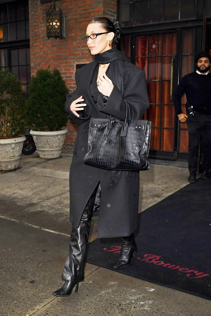 Phong cách all-black luôn là lựa chọn hoàn hảo cho mùa đông. Bella Hadid sử dụng các lớp trang phục với chất liệu đa dạng như áo lụa, quần da bó và áo khoác len bóng để tạo chiều sâu. Cô kết hợp với túi Saint Laurent vân cá sấu và khuyên tai tròn vàng, mang đến vẻ ngoài vừa thanh lịch vừa nổi bật. 