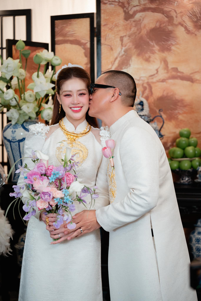 Bộ áo dài được chế tác trên nền vải cao cấp với hiệu ứng ánh kim và tông màu kem sang trọng. Để tạo dấu ấn riêng, nhà thiết kế khéo léo nhấn nhá phần tay áo của cô dâu bằng kiểu dáng tay phồng, đính hoa mềm mại trên vai, cùng tông với hoa văn được đính trên khăn đóng trong suốt của cô dâu.