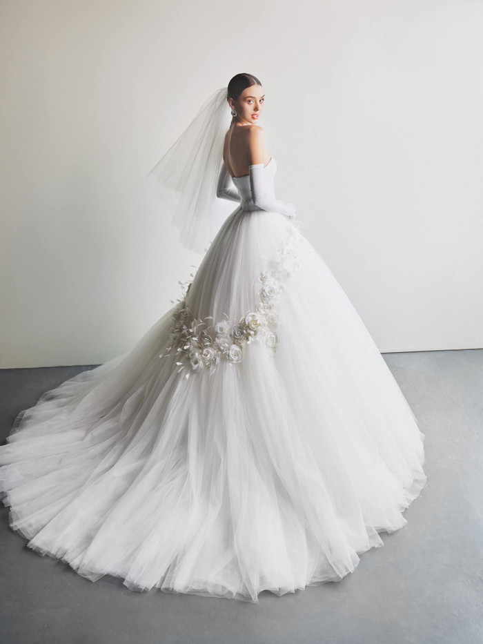 Dianka Zakhidova diện mẫu váy cưới cúp ngực, phần tùng váy lớn, rộng dài quét đất. Thiết kế này còn được tạo điểm nhấn bởi phần hoạ tiết dải hoa hồng, bao tay cùng khăn cài tóc đồng màu. Điểm nhấn của trang phục còn nằm ở phần siết chặt vòng 2, tôn lên triệt để vóc dáng 'đồng hồ cát' của vợ Bùi Tiến Dũng.