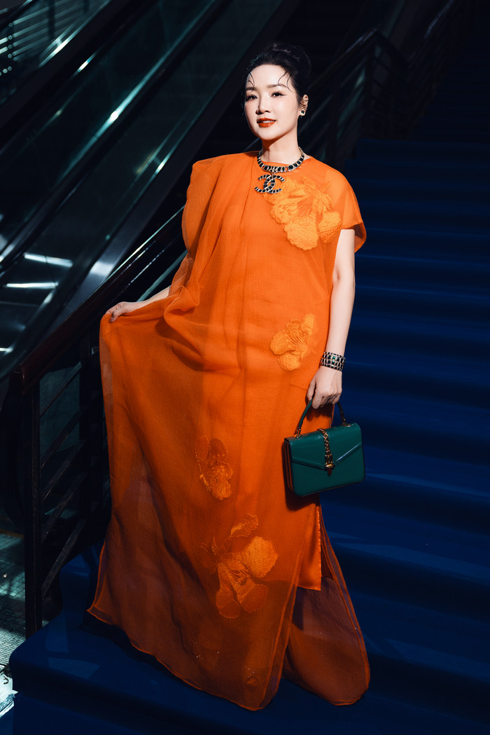 Hoa hậu Đền Hùng khoe khí chất sang trọng trong mẫu đầm cam rộng dáng được làm bằng chất liệu cao cấp có họa tiết chủ đạo hoa phượng. 'Người đẹp không tuổi' phối thêm vòng cổ có ký tự Chanel màu xanh lục bảo 'ton-sur-ton' với túi cầm tay. 
