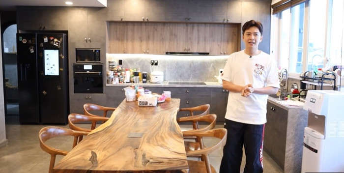 Không gian bếp bài trí đơn giản, sử dụng tông màu trầm. 'Tôi đầu tư một bộ bàn ăn dài đắt tiền để ăn uống cùng gia đình. Tôi rất thích những bữa ăn gia đình nên làm hẳn bàn 8 người rồi mời ba má qua ăn chung để mọi người cùng trò chuyện với nhau. Vì yêu quý gia đình nên tôi dùng tủ lạnh hiện đại, có màn hình trên cánh tủ hiện ảnh gia đình tôi luôn. Tủ lạnh nhà tôi còn có camera bên trong, bấm một cái là coi được đồ ăn qua màn hình, không cần mở ra. Tủ lạnh này còn có cả chục triệu công thức làm đồ ăn để nấu nướng được luôn. Máy giặt nhà tôi cũng xịn, đắt tiền, có chế độ giặt tự động thông minh, tự phân tích mọi thứ để giặt, chỉ cần bấm nút. Tôi chỉ cần bỏ nước giặt một lần vào rồi máy tự dùng cả tháng', Ngô Kiến Huy chia sẻ.