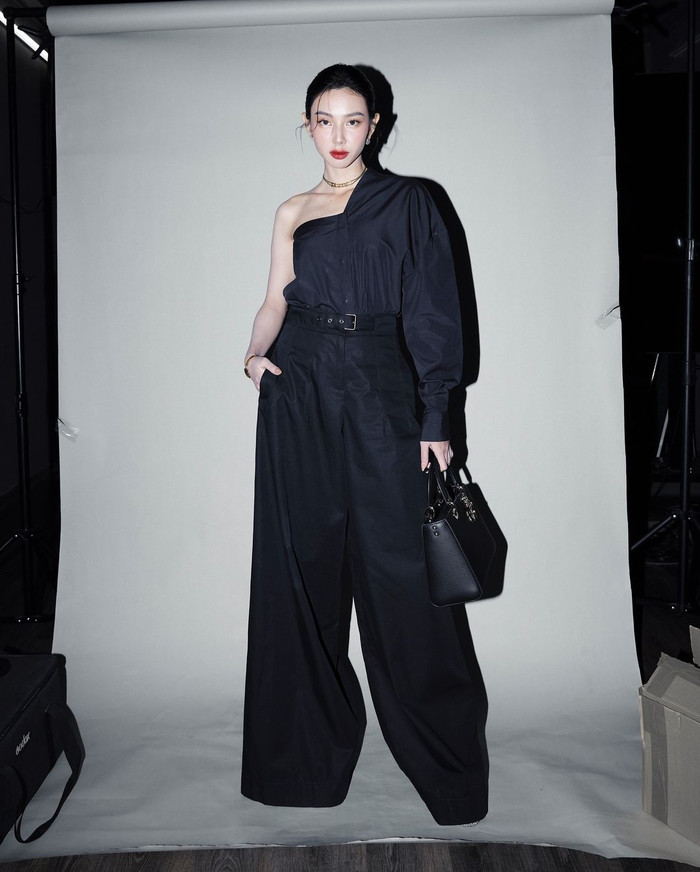 Hoa hậu Thùy Tiên cũng chọn jumpsuit kiểu lệch vai dáng sơ mi trong bộ ảnh gần đây. Thiết kế màu đen làm tôn vinh làn da của Thùy Tiên, phần ống quần dài qua mắt cá khiến Thùy Tiên dễ dàng 'hack chân'. Cô nàng đeo thêm túi cỡ trung, layout của nàng hậu phù hợp trở thành gợi ý cho những chị em công sở. 