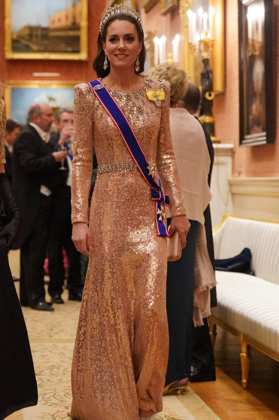 Trong bữa tiệc chiêu đãi của Đoàn ngoại giao tại Cung điện Buckingham, Công nương Kate gây ấn tượng khi tái sử dụng chiếc váy dạ hội màu hồng ánh lấp lánh của Jenny Packham. Nàng phối hợp hoàn hảo cùng chiếc vương miện Lover's Knot Tiara quen thuộc, tạo nên vẻ đẹp vừa hoài cổ vừa kiêu sa.