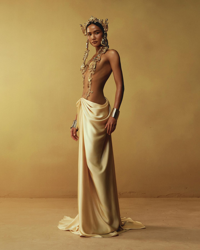 Tông màu chủ đạo là vàng ánh kim và trắng kem, mang đến vẻ đẹp cổ điển nhưng đầy tinh tế. Chất liệu chính sử dụng là lụa cao cấp, giúp tăng thêm độ mềm mại và bắt sáng, làm nổi bật các chi tiết thiết kế. Kỹ thuật in 3D hiện đại được kết hợp cùng kỹ thuật draping thủ công, tái hiện hoàn hảo các họa tiết hoa lá cách điệu và biểu tượng mặt trời.