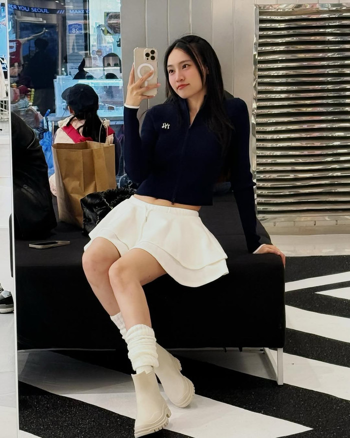 Trong selfie tại trung tâm thương mại, Ninh Dương Lan Ngọc chọn zip-up navy có logo NY, váy trắng xòe ngắn layer và boots trắng chunky, kết hợp tất trắng cao đến đầu gối. Đây là cách mix sporty-preppy kinh điển, nơi item váy từ H&M hay Mango được nâng tầm bởi boots từ Dr. Martens, tạo chiều sâu cho outfit đường phố. Ảnh: IGNV