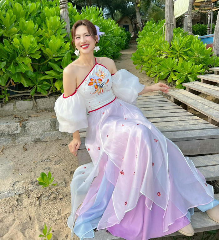 Đôi khi nữ diễn viên đối gió với phong cách nữ tính. Trong bức ảnh ngồi trên lối đi gỗ nhiệt đới, Phương Anh Đào chọn váy dài gradient từ hồng nhạt sang tím và xanh, với phần vai trễ puff sleeve thêu họa tiết chim phượng. Váy có lớp sheer mỏng, tạo hiệu ứng bay bổng, kết hợp giày bệt be đơn giản. Ảnh: FBNV