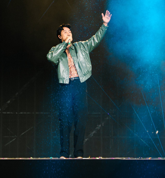 Đen Vâu thường xuyên xuất hiện với các món đồ đơn giản nhưng được phối hợp tinh tế. Trong một buổi biểu diễn, nam ca sĩ chọn áo khoác da màu xanh xám nhạt, chất liệu bóng nhẹ, kiểu dáng bomber cổ điển với viền bo chun ở gấu và tay áo. Bên trong là áo sơ mi họa tiết paisley nhỏ màu cam nâu, cổ bẻ truyền thống, kết hợp quần tây đen thẳng ống và giày thấp cổ tối màu. Ảnh: FBNV