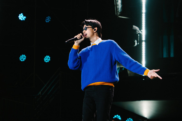 Một outfit khác nổi bật của Đen Vâu là áo len xanh dương royal, dệt kim dày với viền cam contrast ở cổ tay, gấu áo và cổ tròn. Áo len suông rộng, layer trên áo sơ mi trắng lộ phần cổ, phối cùng quần đen ôm và kính gọng tròn. Viền cam mang đến điểm nhấn màu sắc tinh tế, phù hợp với không gian sân khấu tối, giúp Đen Vâu nổi bật mà không cần phụ kiện cầu kỳ. Ảnh: FBNV