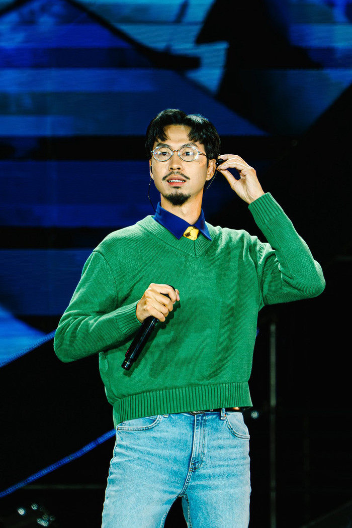 Đen Vâu còn sở hữu chiếc áo len xanh lá cây cổ chữ V. Nam ca sĩ layer áo sơ mi xanh dương bên trong, cà vạt vàng nhỏ và quần jeans xanh nhạt phai. Áo len dệt mịn, cổ V lộ phần cà vạt tạo chiều sâu, trong khi jeans thẳng ống giúp tổng thể casual hơn. Ảnh: FBNV