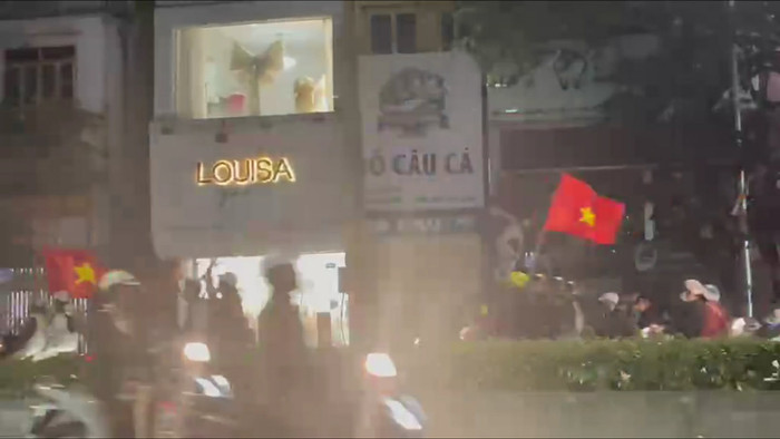 Tại Hà Nội, từ khoảng 22h, dòng người đông đúc xuất hiện trên các tuyến phố chính như Kim Mã, Cầu Diễn - Hồ Tùng Mậu và khu vực gần sân vận động Mỹ Đình. Nhiều người dân tranh thủ bày bán cờ Tổ quốc, băng rôn, kèn trống và áo đỏ sao vàng, tạo nên khung cảnh rực rỡ sắc màu trong dịp SEA Games năm nay. Dù đông đúc, các đoàn xe 'đi bão' vẫn chấp hành tốt quy định giao thông, dừng đèn đỏ nghiêm túc, xếp hàng thẳng tắp tại các nút giao, ghi điểm về ý thức cộng đồng.