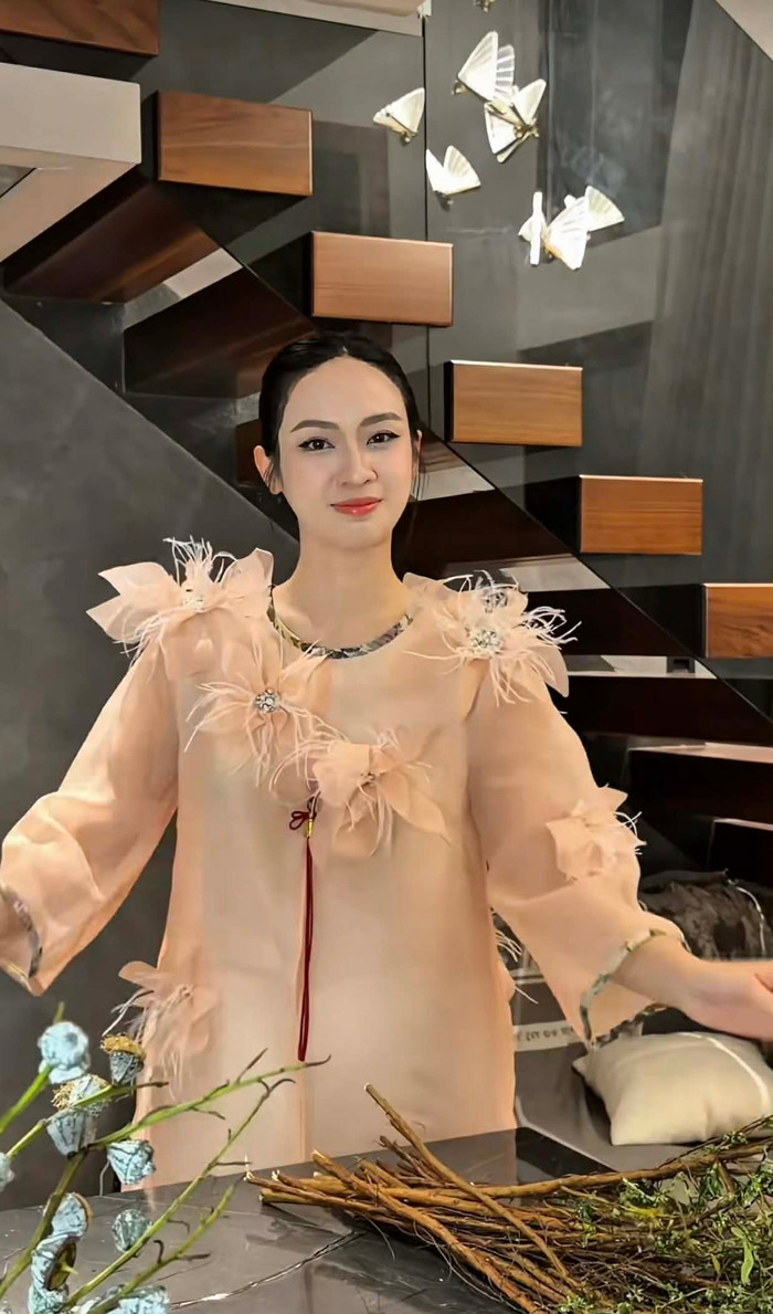 Trong video mới nhất, Á hậu Thúy Quỳnh chọn chiếc áo dài cách tân chất liệu organza màu nude hồng phấn, kiểu dáng dài tay, form rộng rãi nhưng vẫn ôm nhẹ lấy đường nét cơ thể. Điểm nhấn nổi bật nằm ở phần vai và cổ áo được đính kết những bông hoa lớn làm từ lông vũ và vải sheer, tạo hiệu ứng bay bổng, lãng mạn. Chất liệu organza mỏng nhẹ, trong suốt một phần, giúp bộ trang phục toát lên vẻ nhẹ nhàng, nữ tính. Ảnh chụp màn hình