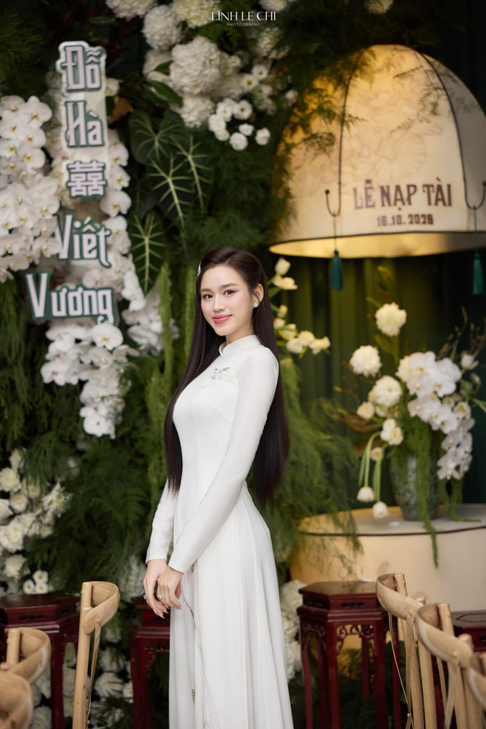 Tại lễ ăn hỏi vào tháng 10/2025, Đỗ Thị Hà diện áo dài trắng kem trong thiết kế mang tên 'Thanh Bình hỷ lạc', thêu tay thủ công với cảm hứng từ trống đồng Đông Sơn, chim Lạc Việt, và sóng biển, tượng trưng cho quê hương và sự gắn kết lứa đôi. Bộ áo dài này sử dụng lụa Nhật, nhấn nhá họa tiết hoa sen và kim tuyến lấp lánh, tạo hiệu ứng trong suốt nhẹ nhàng, tôn lên vóc dáng mảnh mai. Nàng hậu phối cùng giày thấp đơn giản, tránh làm nổi bật chiều cao mà tập trung vào sự hài hòa tổng thể. Ảnh: FBNV