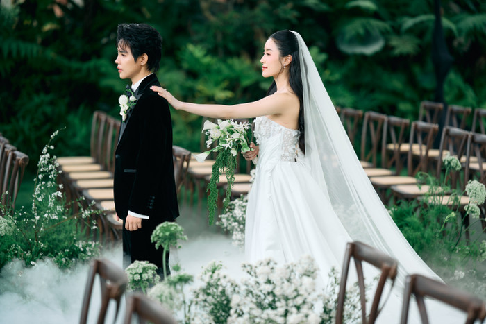 Vũ Cát Tường trong bộ vest nhung đen lịch lãm lặng người, chờ đợi khoảnh khắc được nhìn thấy người bạn đời của mình. Không gian xung quanh phủ đầy sắc xanh và những bông hoa trắng tinh khiết, tạo nên một khung cảnh lãng mạn.