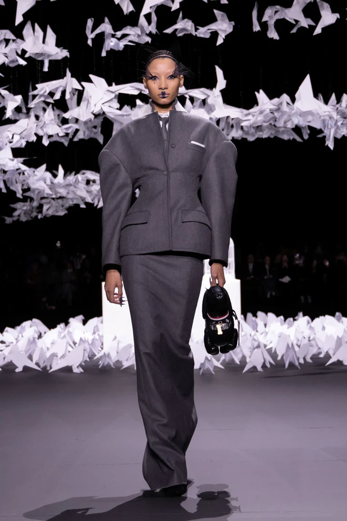 Mở màn show diễn với thiết kế suit màu xám mang tính biểu tượng của nhà mốt với phần vai độn phồng tràn xuống cùng điểm nhấn chiết eo sâu kết hợp với chiếc túi được thiết kế một cách kì lạ mang logo sọc đỏ trắng xanh của Thom Browne. (Ảnh: Thom Browne Fall 2025)
