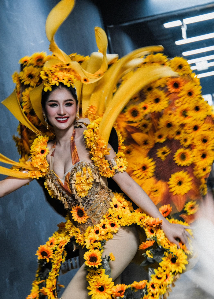 Thí sinh khác diện trang phục tái hiện hình ảnh hoa hướng dương. Thiết kế này gây chú ý với phần cánh đeo sau lưng cỡ lớn, đính kết dày đặc loài hoa được nhắc tên. Phần trang phục được người đẹp diện lại mang kiểu dáng jumpsuit liền thân bó sát giúp tôn lên những đường cong nóng bỏng của hình thể. (Ảnh: Miss Grand Thailand)