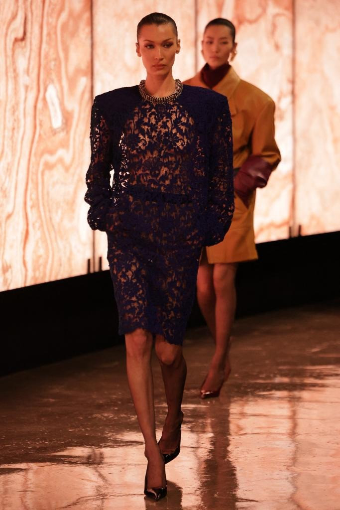 Bella Hadid sải bước trong bộ trang phục ren xuyên thấu màu xanh navy đậm chất haute couture. Thiết kế bao gồm áo dài tay kết hợp với chân váy đồng bộ, tạo nên vẻ ngoài tinh tế nhưng không kém phần táo bạo. Chất liệu ren mỏng manh phủ lên làn da, để lộ những đường nét cơ thể quyến rũ, nhưng vẫn được cân bằng bởi chi tiết cổ áo đính đá lấp lánh và phần cầu vai mở rộng, mang đến cảm giác quyền lực và mạnh mẽ.