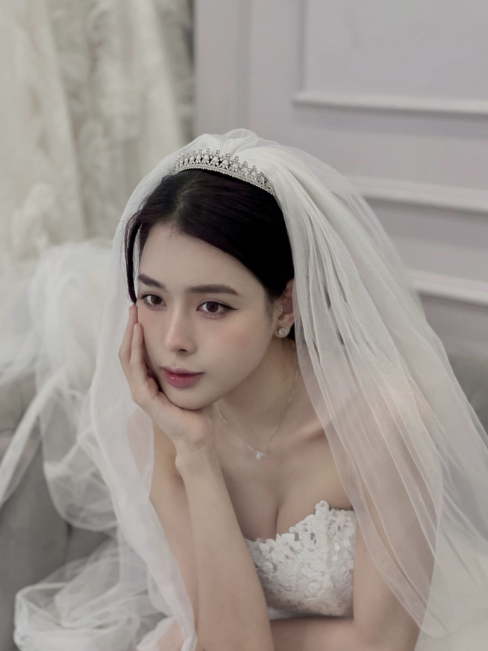 Trong ảnh, bạn gái Vũ Văn Thanh khoác lên mình chiếc váy cưới mang phong cách thanh lịch và tinh tế. Thiết kế cúp ngực phối ren đính hoa tinh xảo giúp tôn lên đường nét quyến rũ của cô dâu, đồng thời tạo điểm nhấn mềm mại, nữ tính. Chiếc khăn voan dài thướt tha kết hợp cùng vương miện lấp lánh mang lại vẻ đẹp cổ điển, sang trọng cho Bích Hạnh. Gam màu trắng truyền thống không chỉ tôn da mà còn thể hiện sự thuần khiết, làm nổi bật nét đẹp của cô dâu trong ngày trọng đại.