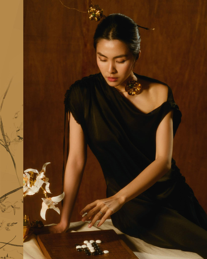 Nữ diễn viên hóa thân thành một quý cô cổ điển với chiếc đầm đen cắt xẻ tinh tế. Bộ trang phục có phom dáng bất đối xứng, kết hợp cùng phụ kiện ánh vàng tạo nên vẻ ngoài huyền bí. Với ánh mắt trầm tư và bàn cờ vây trước mặt, hình ảnh của cô gợi lên sự sâu lắng đầy nghệ thuật.