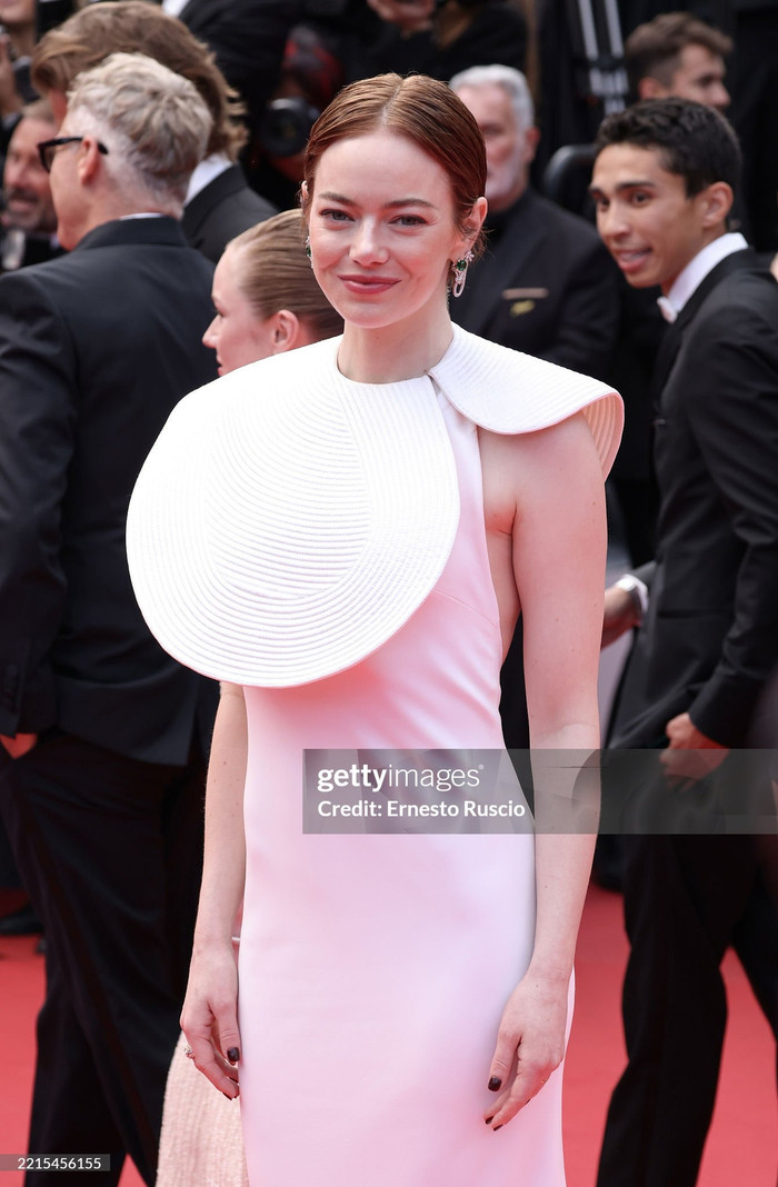 Emma Stone chọn cho mình một thiết kế đầy thách thức thị giác từ Louis Vuitton, thương hiệu mà cô là gương mặt đại diện lâu năm. Chiếc đầm hồng phấn ôm sát cơ thể với chất liệu satin cao cấp, nhưng điểm nhấn gây bùng nổ chính là phần cổ áo tạo hình xoắn ốc 3D bất đối xứng như một tác phẩm điêu khắc hiện đại, trùm lên toàn bộ vai phải.