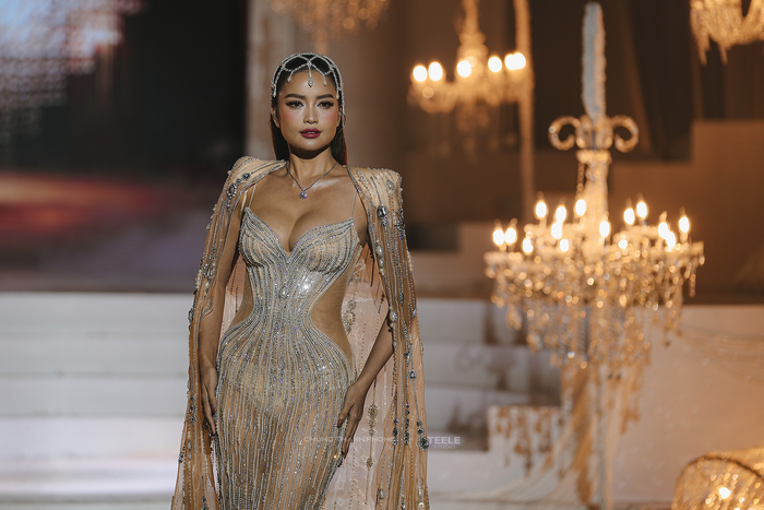 Bộ cánh của Ngọc Châu trong bộ sưu tập thực sự là một sự giao thoa giữa vẻ lộng lẫy của một cô dâu và thần thái quyền lực của một beauty queen. Thân váy được thiết kế ôm sát, tôn vinh trọn vẹn đường cong quyến rũ của một hoa hậu. Trên nền vải xuyên thấu màu nude, hàng ngàn viên đá, pha lê và ngọc trai được đính kết thủ công theo những đường cong uốn lượn, tạo hiệu ứng thị giác như ánh sáng bao bọc cơ thể. Những khoảng hở (cut-out) táo bạo và đối xứng hai bên eo không chỉ tăng thêm vẻ gợi cảm mà còn là một chi tiết quen thuộc trong các thiết kế dạ hội của các hoa - á hậu, giúp khoe khéo vòng eo thon gọn.