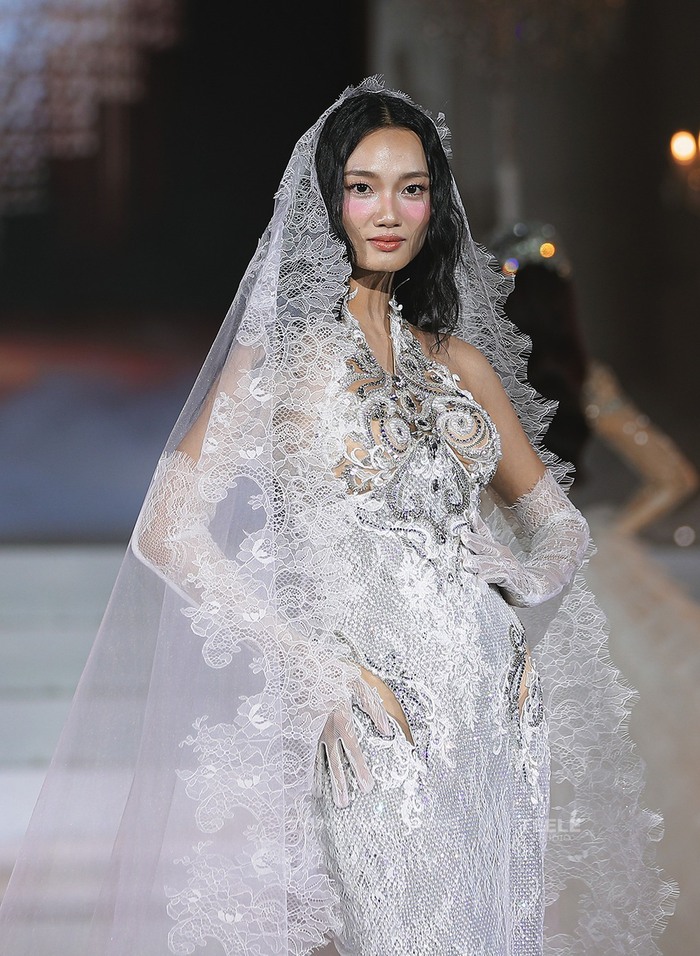 Á hậu Quỳnh Anh mang đến hình ảnh một cô dâu mới lạ, đầy quyến rũ và tinh xảo trong bộ cánh lộng lẫy. Điểm nhấn chủ đạo là chiếc khăn voan trùm đầu được chế tác từ chất liệu ren cao cấp, với những họa tiết hoa văn thêu nổi vô cùng tinh tế và đường viền đăng-ten mềm mại, buông dài thướt tha, gợi lên nét e ấp và thanh lịch truyền thống. Thân váy ôm sát, dù được bao phủ bởi hàng ngàn viên đá và pha lê lấp lánh, vẫn khéo léo lồng ghép các chi tiết ren đồng điệu ở phần cổ yếm cách điệu và những đường viền cut-out táo bạo ở hông, tạo sự cân bằng giữa vẻ đẹp cổ điển và nét gợi cảm hiện đại.