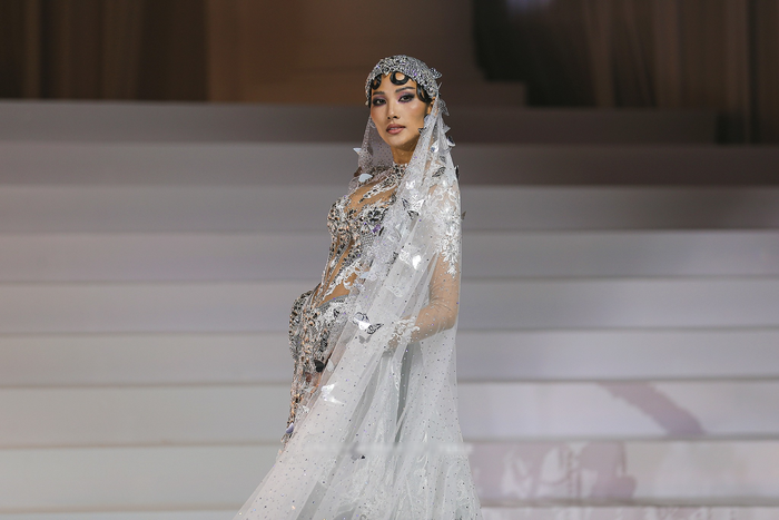 Đúng với tinh thần 'xuất hiện cuối cùng', Hoàng Thùy khiến toàn bộ khán phòng phải 'wow' khi khoác trên mình thiết kế đậm chất haute couture. Bộ cánh toát lên vẻ lộng lẫy, kiêu sa và đầy huyền ảo. Điểm nhấn nổi bật là chiếc mũ đội đầu và khăn trùm được đính kết đá quý, pha lê tinh xảo, kết hợp với khăn voan mỏng buông dài. Chiếc veil điểm xuyết hàng trăm cánh bướm kim loại lấp lánh như một đàn bướm sống động.   