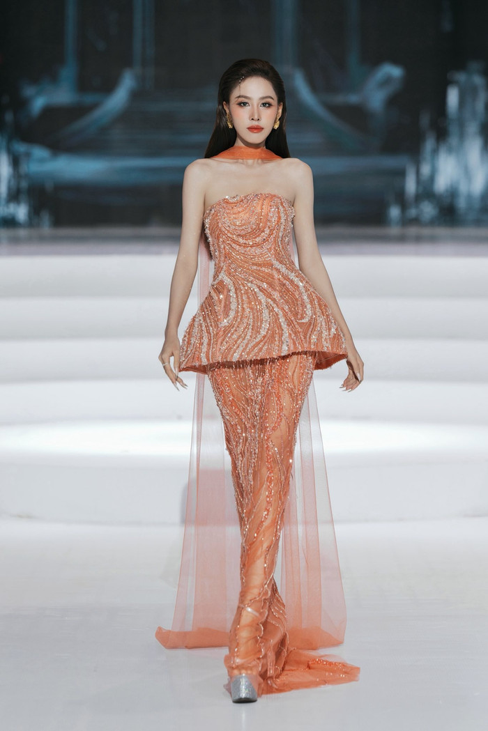 Á hậu Ngọc Hằng nổi bật trong thiết kế dạ hội màu cam ánh kim, được xử lý kỹ thuật đính kết kỳ công tạo hiệu ứng lấp lánh sống động. Dáng váy ôm sát tôn dáng, phối cùng chi tiết peplum tạo phom lạ mắt và tà voan mỏng rũ nhẹ phía sau. Tổng thể mang đến vẻ kiêu sa, sắc sảo nhưng không kém phần bay bổng.