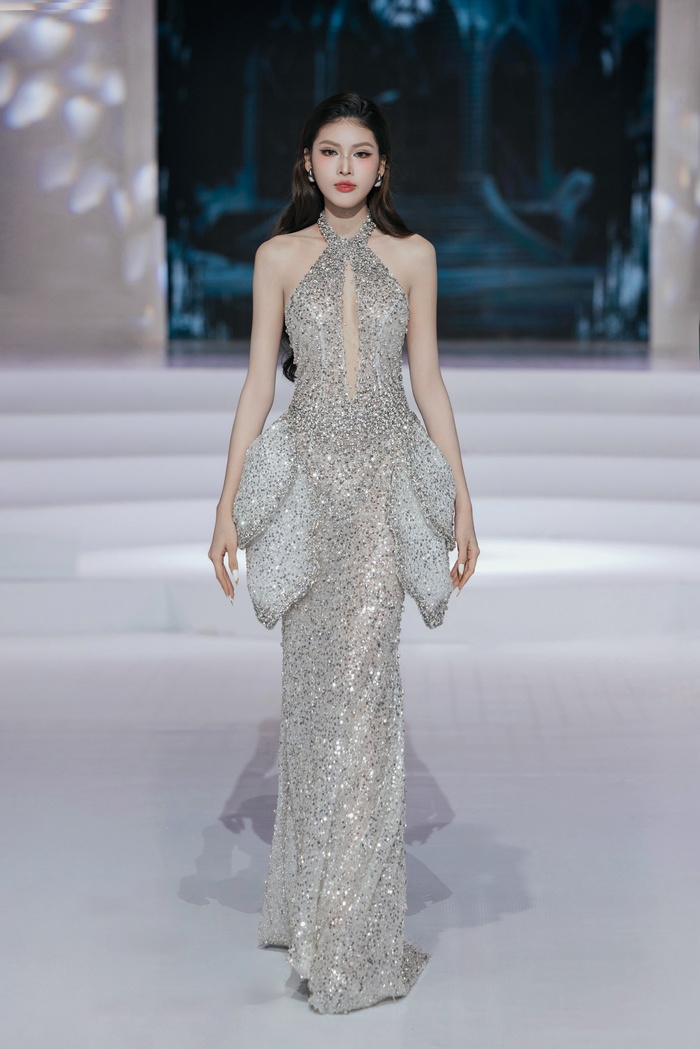 Á hậu Ngọc Thảo khoe trọn vóc dáng trong thiết kế dạ hội ánh bạc ôm sát, phủ kín sequins lấp lánh. Chi tiết cổ yếm khoét ngực sâu tạo điểm nhấn quyến rũ, kết hợp phần hông xếp lớp cấu trúc mềm mại như cánh hoa nở. Tổng thể vừa hiện đại, vừa gợi cảm, giúp cô tỏa sáng với phong thái tự tin và kiêu hãnh.