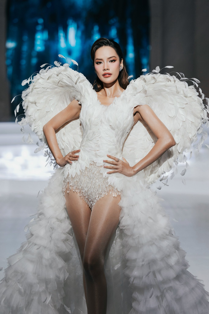 Hoa hậu Lê Hoàng Phương diện thiết kế bodysuit trắng ôm sát, phủ lông vũ cao cấp và pha lê lấp lánh. Điểm nhấn là đôi cánh khổng lồ dựng hình trái tim, tạo hiệu ứng thiên thần quyền lực. Phần đuôi váy tulle xếp tầng bay bổng, kết hợp makeup sắc sảo và tóc uốn nhẹ, tôn trọn thần thái first-face. 