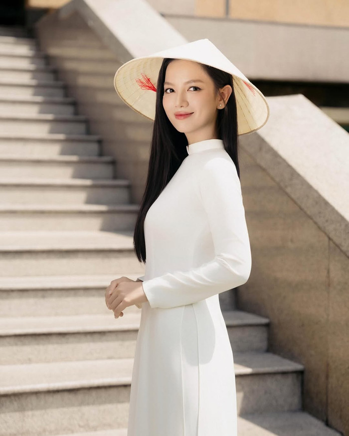 Lê Hạ Anh thường xuất hiện với nhiều phiên bản áo dài đa dạng, từ thiết kế cổ điển đến cách tân tinh tế. Một hình ảnh nổi bật là tà áo dài trắng tinh khôi, chất liệu lụa mỏng nhẹ ôm sát cơ thể, tôn lên vóc dáng mảnh mai và làn da sáng mịn. Chiếc nón lá trắng truyền thống được điểm xuyết bằng chi tiết đỏ rực, tạo điểm nhấn dân dã nhưng thanh lịch, gợi nhớ hình ảnh người phụ nữ Việt Nam trong không gian đô thị cổ kính với cầu thang dài dẫn lên. Tóc dài buông xõa tự nhiên, makeup nhẹ nhàng nhấn vào đôi mắt to và nụ cười mỉm, mang lại cảm giác gần gũi, trong trẻo. Ảnh: IGNV