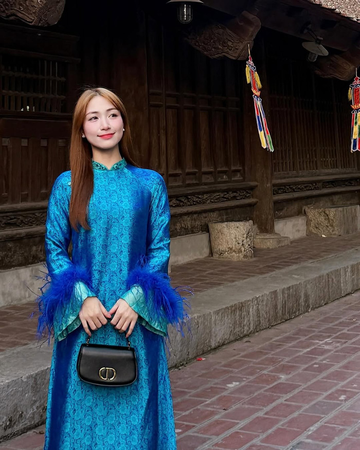 Tiếp theo là mẫu áo dài màu xanh dương đậm của Hòa Minzy, được may từ vải brocade với hoa văn tinh tế dệt nổi. Điểm nhấn nằm ở phần tay áo loe rộng, được trang trí thêm lớp lông vũ gradient từ xanh nhạt đến xanh đậm, tạo hiệu ứng chuyển động mềm mại mỗi khi di chuyển. Phần thân áo dài được cắt may ôm vừa vặn, độ dài chạm gót, mang lại vẻ ngoài sang trọng nhưng không quá cứng nhắc. Mẫu áo dài này thể hiện cách Hòa Minzy biến tấu yếu tố truyền thống bằng chi tiết cách tân hiện đại, phù hợp cho các sự kiện Tết ngoài trời hoặc những buổi gặp gỡ trang trọng. Ảnh: IGNV