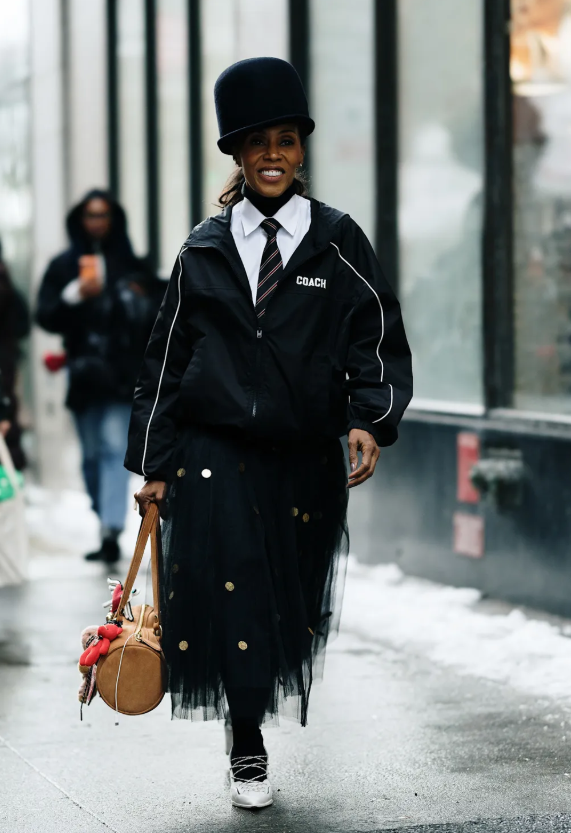 Phong cách thể thao (sportif style) trở thành xu hướng nổi bật năm 2026, kết hợp giữa trang phục thể thao và thiết kế thanh lịch tưởng chừng đối lập. Stylist June Ambrose tạo điểm nhấn khi phối áo khoác gió cùng chân váy voan đính sequin, hoàn thiện bằng cà vạt thắt gọn trên nền sơ mi trắng. Ảnh: Launchmetrics