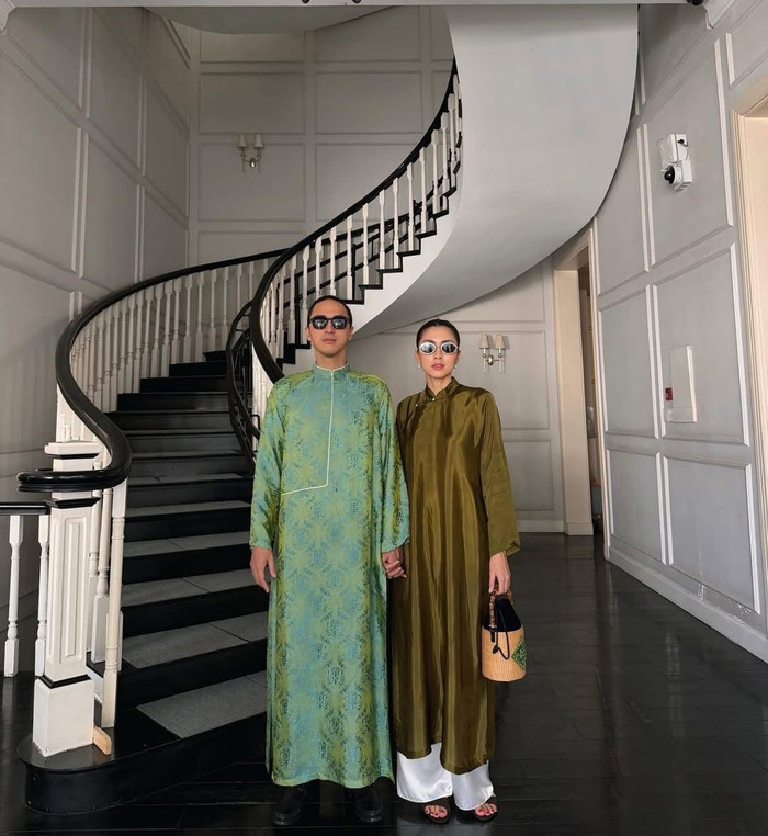 Doanh nhân Louis Nguyễn diện áo dài xanh lá nhạt in họa tiết thư pháp chìm, form suông rộng, cổ cao truyền thống tạo vẻ điềm đạm, cổ điển. Tăng Thanh Hà mặc áo dài lụa satin màu olive ánh nhẹ, thiết kế tối giản, kết hợp quần trắng và túi cói nhỏ, tổng thể vừa hiện đại vừa sang trọng. Ảnh: FBNV