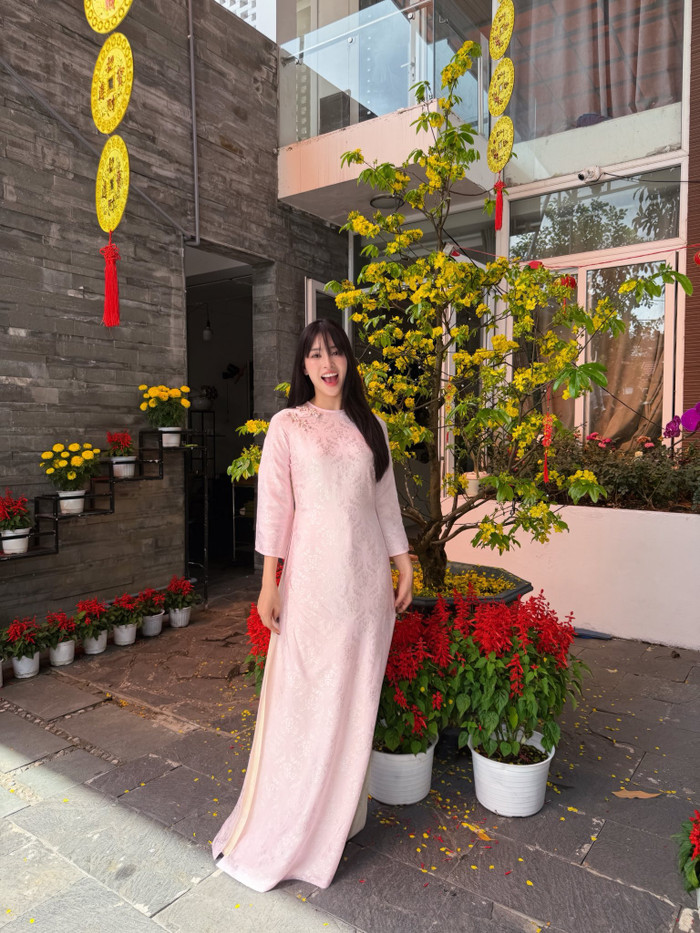 Hoa hậu Tiểu Vy dịu dàng trong tà áo dài hồng phấn với họa tiết dập chìm nhẹ trên nền vải, tay lỡ và cổ cao truyền thống. Tổng thể mang lại cảm giác dịu dàng, thanh lịch, đặc biệt nổi bật khi đứng cạnh mai vàng và hoa xuân rực rỡ. Ảnh: FBNV