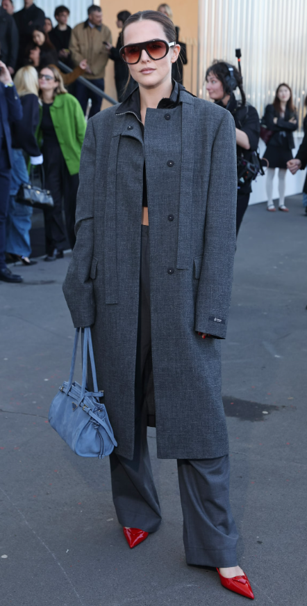 Zoey Deutch diện bộ suit xám phom rộng với áo khoác dáng dài, phối cùng kính râm bản lớn tạo vẻ ngoài quyền lực. Điểm nhấn là đôi giày mũi nhọn đỏ nổi bật, giúp tổng thể trung tính thêm phần cá tính. Ảnh: Getty Images