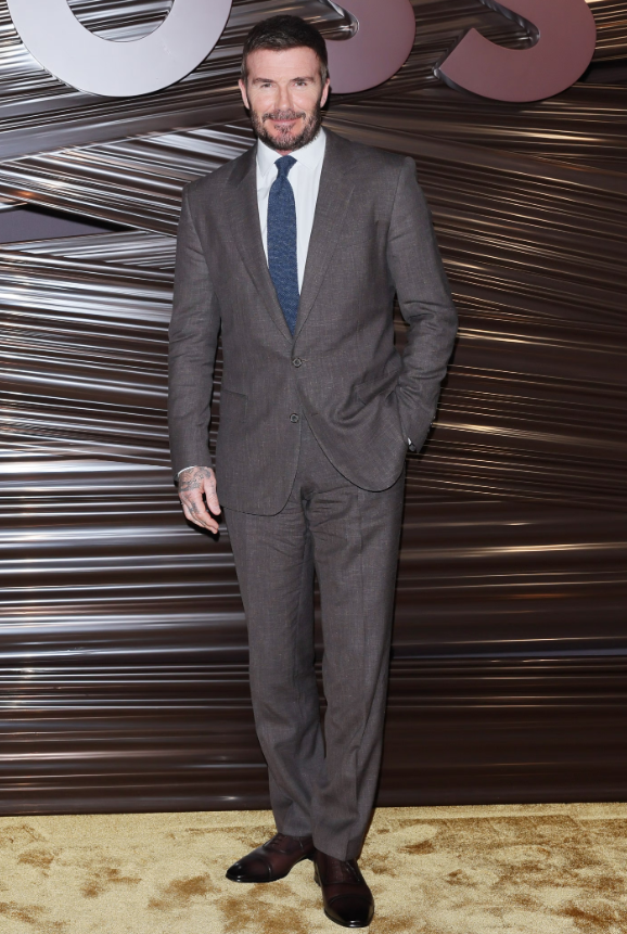 David Beckham xuất hiện với bộ suit nâu chuẩn mực phối cùng sơ mi trắng và cà vạt xanh, mang đến vẻ lịch lãm cổ điển. Phom dáng vừa vặn tôn lên phong thái chững chạc, sang trọng. Ảnh: Getty Images