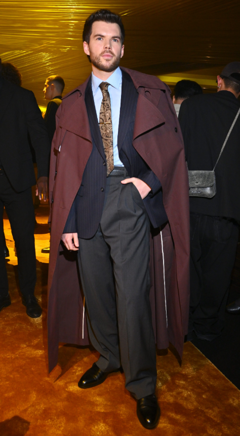 Luke Newton phối bộ suit kẻ sọc với sơ mi xanh nhạt và cà vạt họa tiết tạo cảm giác hiện đại, chỉn chu. Áo trench coat màu rượu vang khoác ngoài giúp tổng thể thêm chiều sâu và ấn tượng. Ảnh: Getty Images