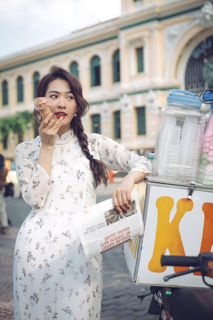 Không chỉ gắn với vẻ đẹp cổ điển, Minh Trang còn mang đến cảm giác gần gũi, đời thường khi diện áo dài trắng in hoa nhí. Phụ kiện ngọc trai kết hợp bối cảnh xe kem ven đường tạo nên tổng thể vừa trẻ trung, vừa hoài cổ, gợi nét duyên ngầm rất riêng. Ảnh: FBNV