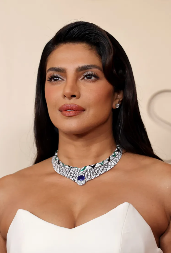 Priyanka Chopra mang đến vẻ đẹp sang trọng với mái tóc dài óng ả chạm eo. Nhà tạo mẫu tóc Bridget Brager tạo điểm nhấn bằng một lọn sóng lớn được tạo hình cao trên trán, giúp tôn lên độ bóng như gương của mái tóc. Kiểu tóc vuốt gọn ra sau cũng khiến mọi ánh nhìn đổ dồn vào chiếc vòng cổ sapphire nổi bật của Bvlgari.