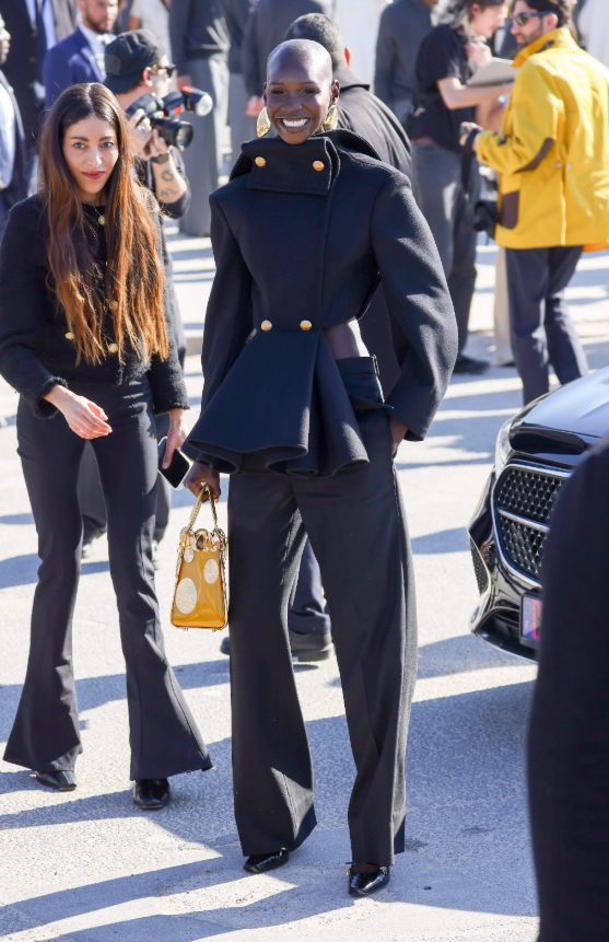 Adot Gak lựa chọn áo khoác đen dáng peplum với phần vai phồng và hàng khuy kim loại nổi bật, phối cùng quần âu ống rộng đồng màu. Túi xách tông vàng tạo điểm nhấn sáng, tôn lên hình ảnh quyền lực và thời thượng. Ảnh: Getty Images