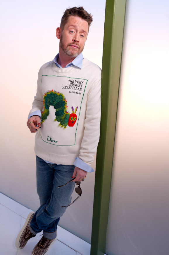 Xuất hiện ở tuần lễ thời trang Paris Thu Đông 2026, Macaulay Culkin mặc áo len màu kem thêu hình nổi bật phía trước, kết hợp quần jeans xanh cơ bản và giày da nâu. Bên trong là áo sơ mi xanh nhạt lộ cổ và tay áo. Trang phục mang tinh thần smart-casual, pha chút trẻ trung nhờ họa tiết sinh động trên áo. Ảnh: Getty Images