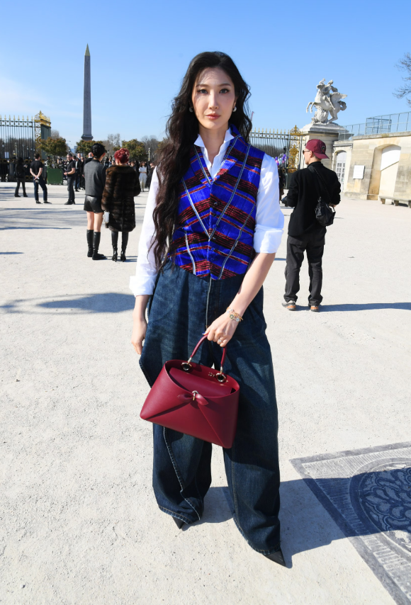 Ejae diện sơ mi trắng kết hợp áo gile kẻ ô xanh - đỏ nổi bật, phối cùng quần jeans ống rộng tối màu. Túi xách đỏ tạo điểm nhấn tương phản, mang đến tổng thể preppy pha street style hiện đại. Ảnh: Getty Images