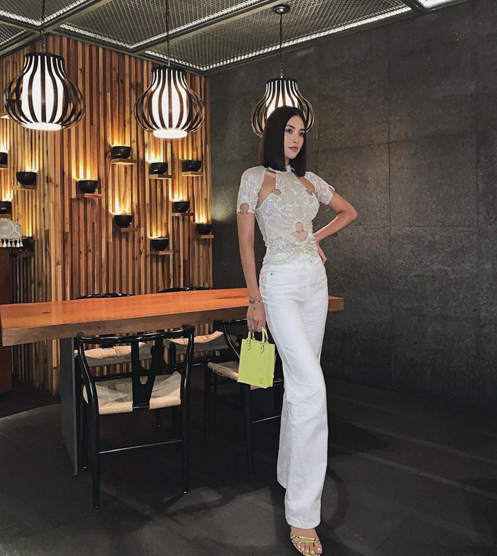 Kiểu áo chất liệu sheer xuyên thấu với những phần cut out trên thân áo được Hoa hậu Tiểu Vy mix cùng quần trắng và phụ kiện túi nhỏ màu xanh lá chuối 