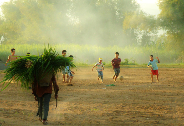 Người đàn ông đi cắt mụt mía (cây mía non) về cho bò ăn, đi ngang qua sân bóng 'dã chiến' của nhóm trẻ em trong thôn. Cứ mỗi chiều, sau khi tan lớp hoặc tạm xong việc nhà, nhóm trẻ và cả người lớn ở thôn Phước Lộc, xã Xuân Quang 3, H.Đồng Xuân lại dợt vài trận banh dưới nắng chiều buông. ẢNH: VÕ TIẾN