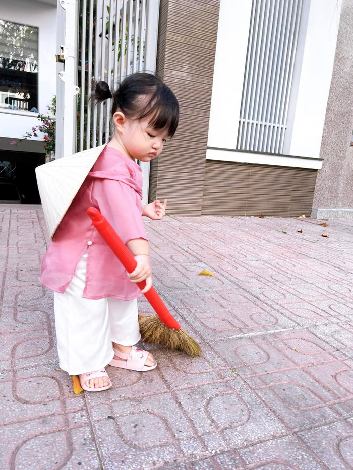 Đông Nhi chia sẻ trên trang Facebook khoảnh khắc con gái diện bộ quần áo bà ba, đội nón, tay cầm chổi quét sân rất chuyên nghiệp.
